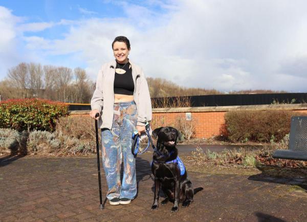 A woman with short black hair and a walking sticks hold a black support dog on a lead and smiles
