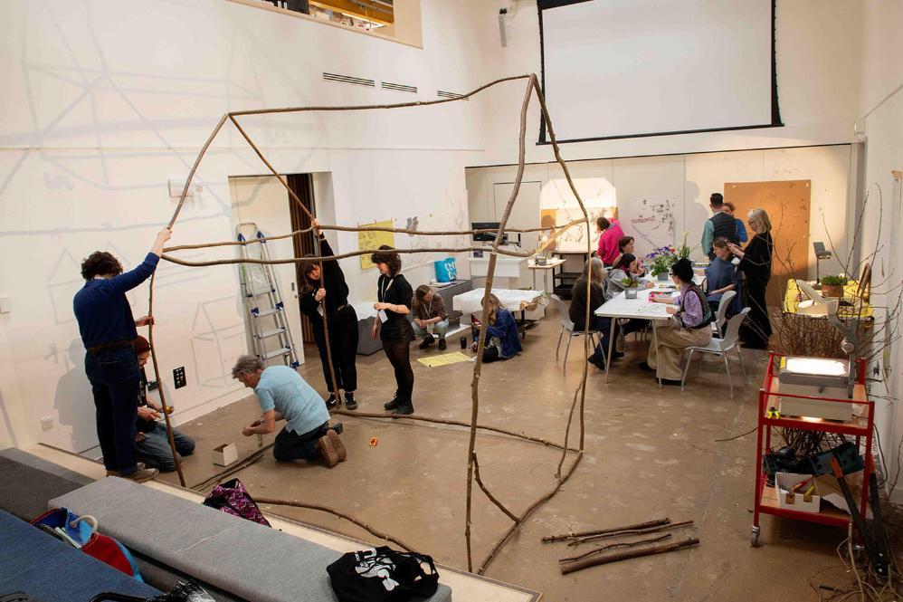 people focus on building a house frame out of wood, more gather around a table in the background.
