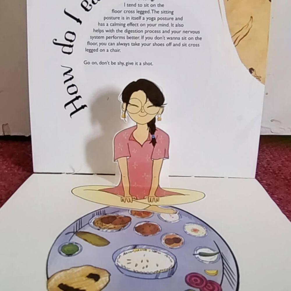 A pop up in a book showing a girl sat crossed legged eating a meal
