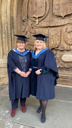 A mother and daughter stand in front of Bath Abbey. They are both in black and blue graduation robes.