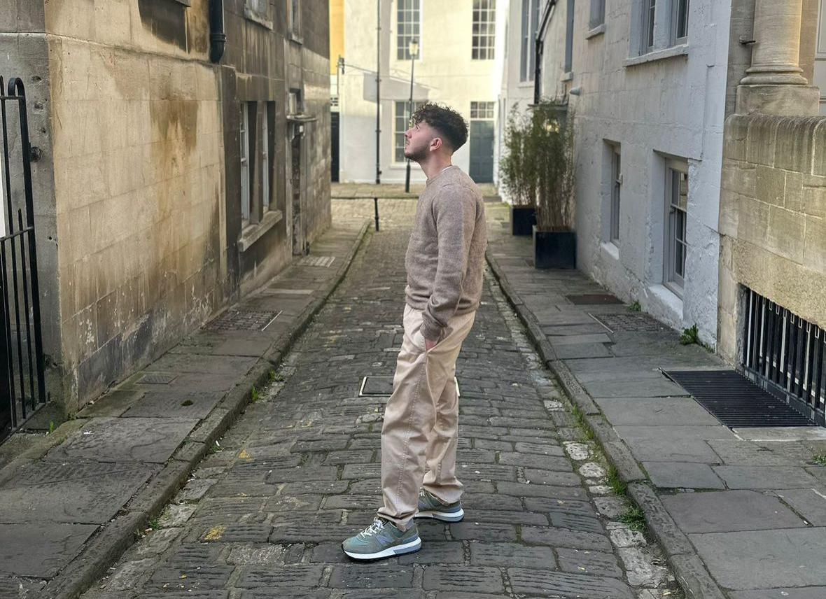 a man stood on a cobbled street in a beige outfit in front of old bath stone buildings