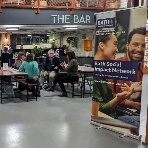 A group of people sat in front of a bar with a large banner displaying the words 'Bath Social Impact Network'