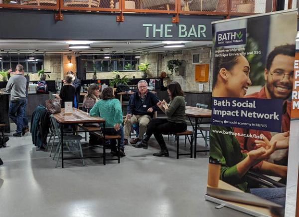 A group of people sat in front of a bar with a large banner displaying the words 'Bath Social Impact Network'