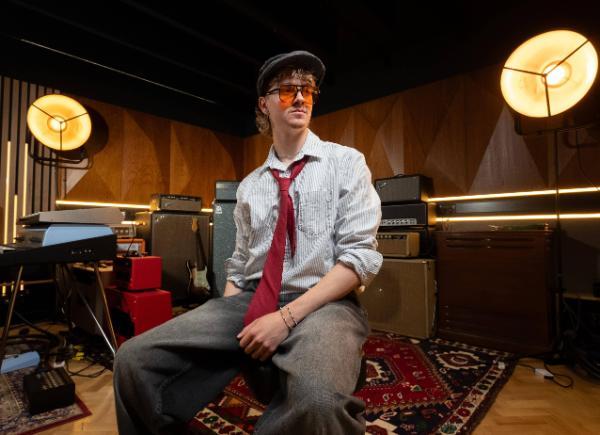 A student wearing a shirt and tie in a music room, with large amp speakers behind him