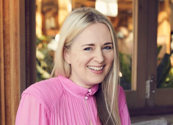 A person with long, blonde hair and wearing a pink dress smiles at the camera