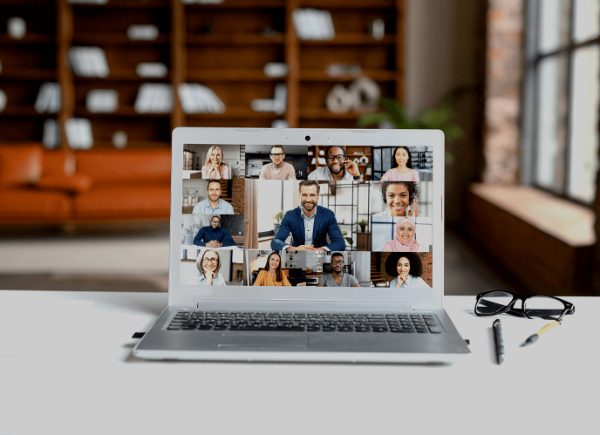 a laptop sits on a table. On the screen are thirteen people of different ethnicities talking on a video call.