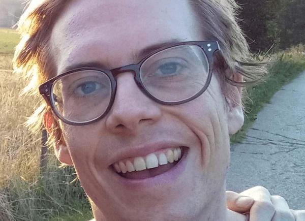 Headshot of someone with short blonde hair and glasses smiling. Hand on shoulder of purple T-shirt.