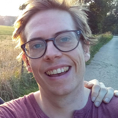 Headshot of someone in glasses and purple t shirt on a country lane