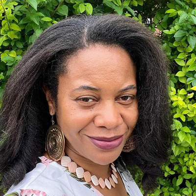 headshot of someone with long dark hair and gold necklace smiling in front of hedge