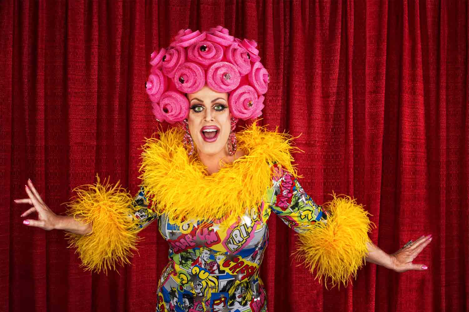 Person with pink swirls for hair and yellow feather boa dressed in drag in front of red curtain