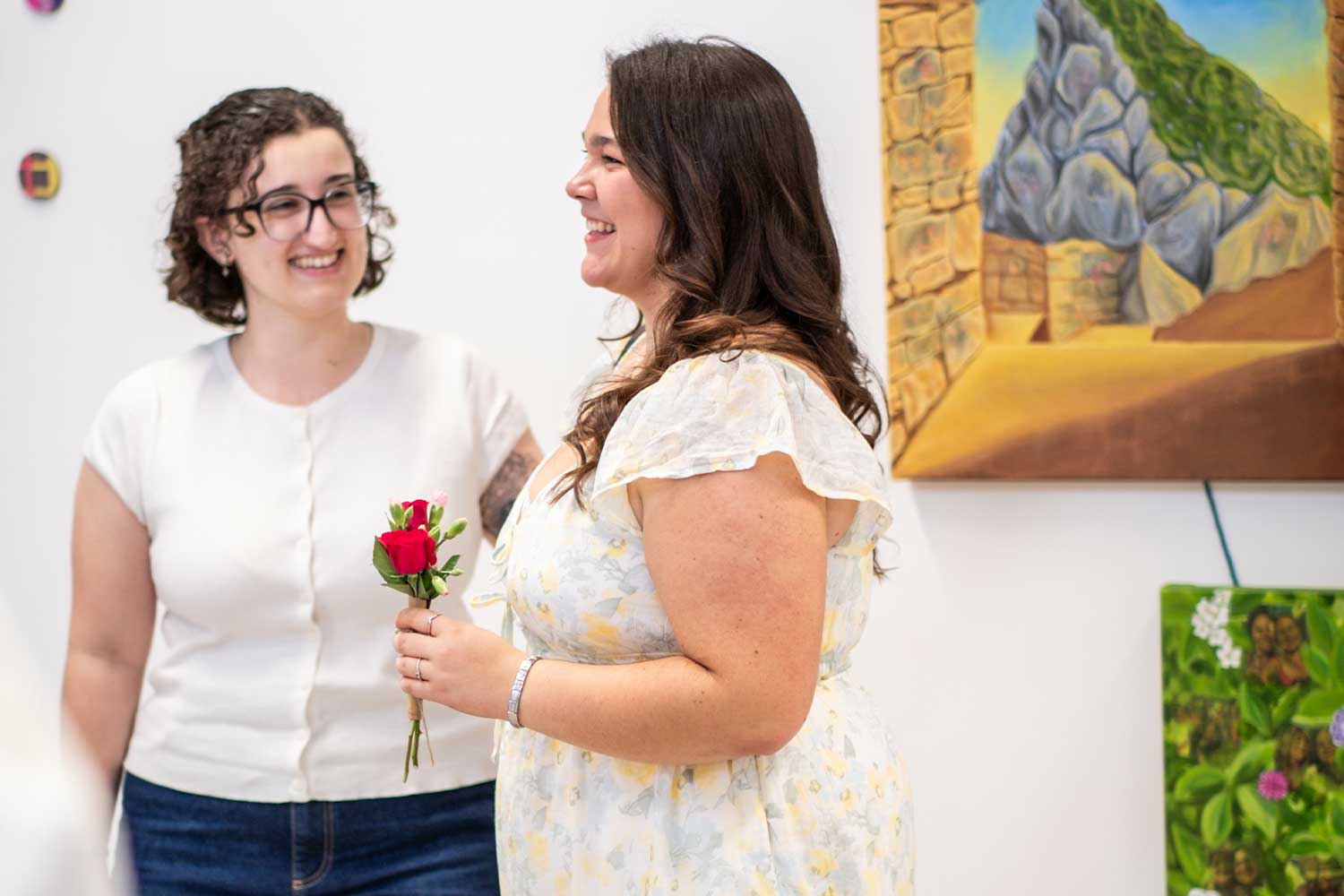 Two people smiling as they stand in front of paintings. One is holding a single red rose.