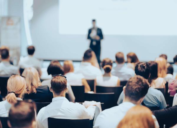 An audience of seated people look at a speaker standing at the front of the room