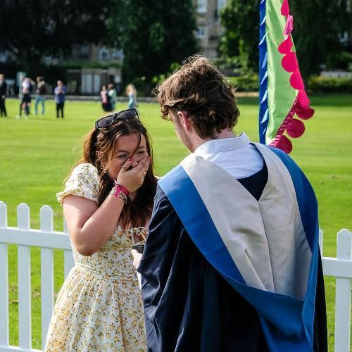 A graduate in ceremonial gown looks towards a woman in a flowery dress who has her hand over her mouth in surprise. They are in a grass area with festival banners and a picket fence behind them.