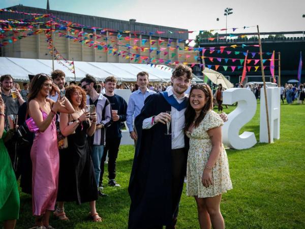 A graduate in ceremonial gown stands next to a woman in a flowery dress smiling. The graduate holds a champagne glass in one hand, the other around her shoulders. They are in a grass area with festival banners and a marquee behind them. People are gathered around them in celebration.