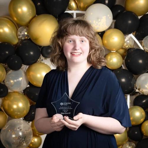 A girl in a smart dress stands in front of a gold balloon arch whilst holding an award