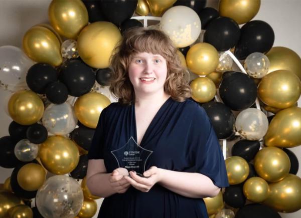 A girl in a smart dress stands in front of a gold balloon arch whilst holding an award