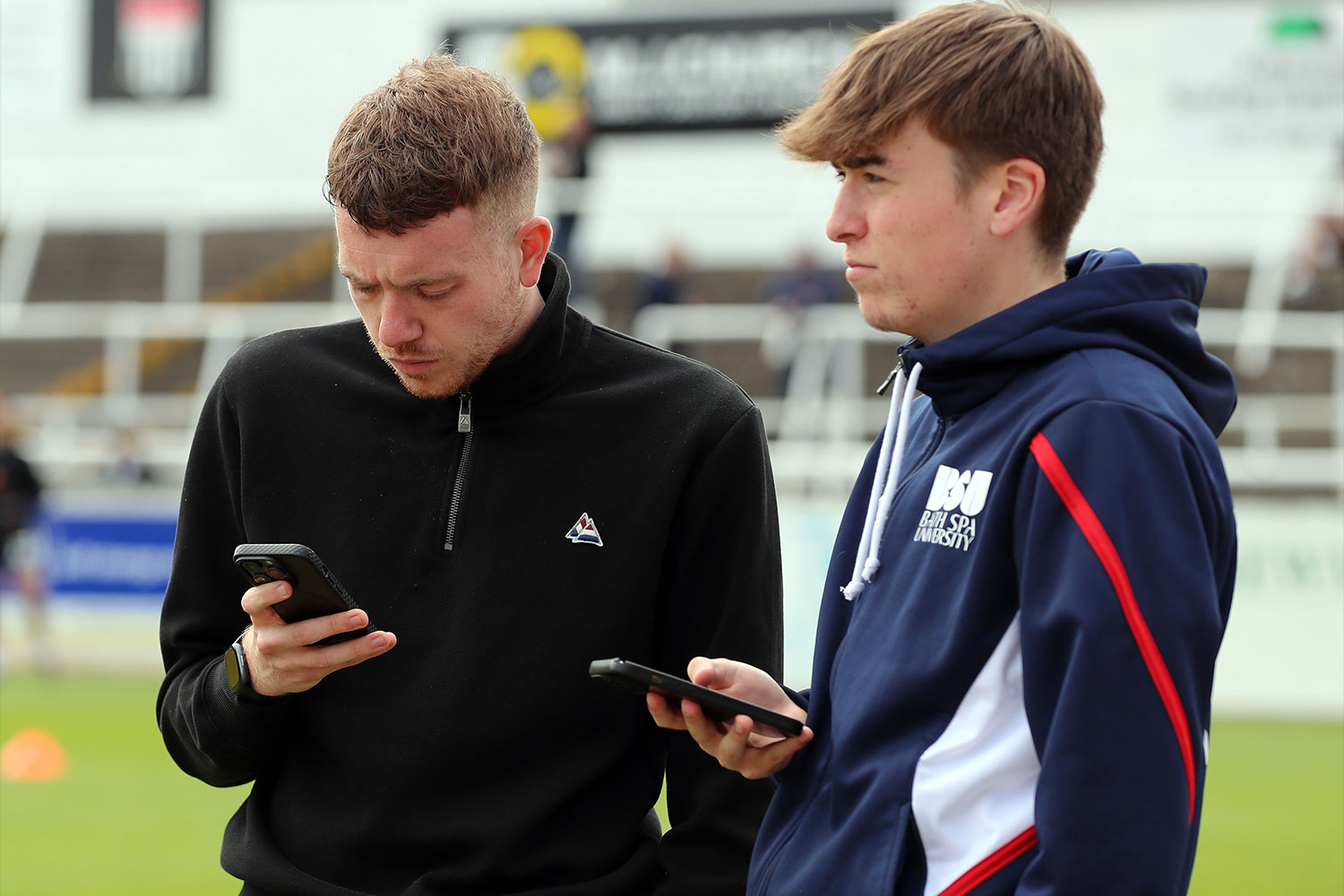 two men looking at phones on a football pitch