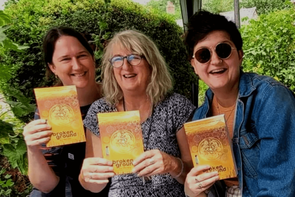 A mother and her two daughters sit in a garden. They all hold up a copy of a book called broken ground.