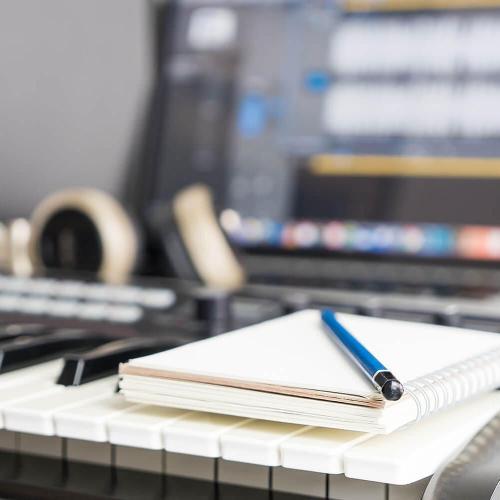A notebook and pencil rest on top of a keyboard. Monitors showing music editing software, and headphones are blurred in the background.