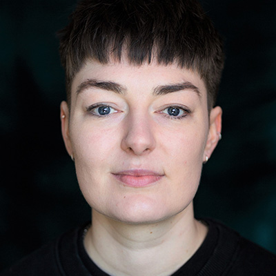 A headshot of CJ Turner-McMullan looking at the camera against a dark background