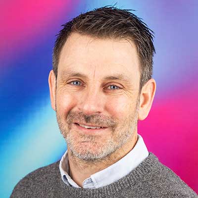 A person with cropped dark hair and a grey beard smiles at the camera