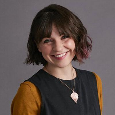 Smiling young woman with brown bob in a grey dress and yellow undershirt.