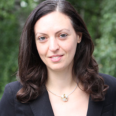 A profile photo of a woman with dark hair wearing a black dress standing in front of trees