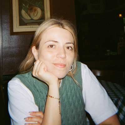 A person with long blonde hair leans on a table and looks at the camera