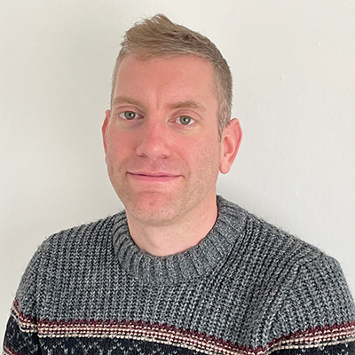 A white man with short blond hair, slightly smiling and wearing a grey jumper.