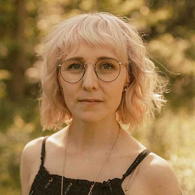 A person with a short, pale pink bob and round glasses looks at the camera