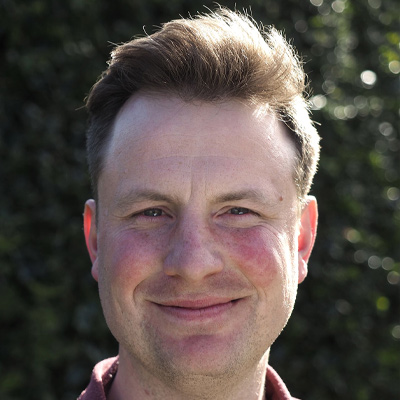 Person with short light brown hair smiling in sunlight in front of green hedging