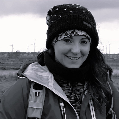 Person in hat with long hair smiling at camera in rural setting with fields behind