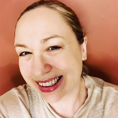 Headshot of person with blonde hair tied back in cream top smiling