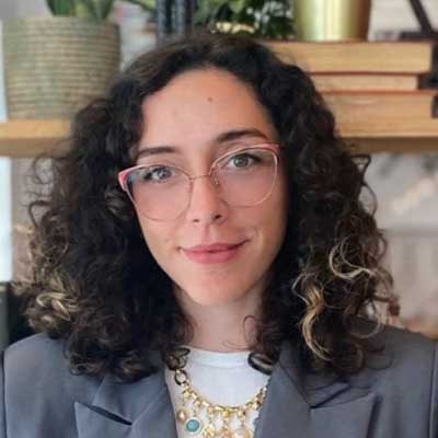 A person with dark, curly hair and pink glasses looks at the camera