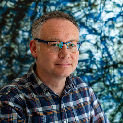 Headshot of person in checked shirt short hair and glasses smiling
