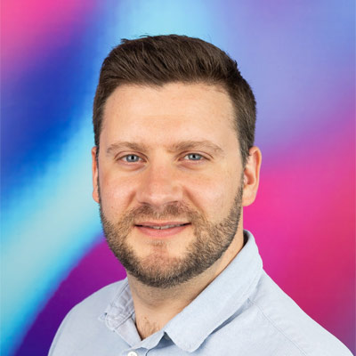 Person with short dark hair and beard in pale shirt smiling at camera
