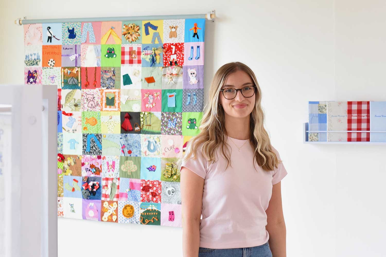 A person with long blonde hair stands in front of a bookshelf and a patchwork quilt hanging on the wall