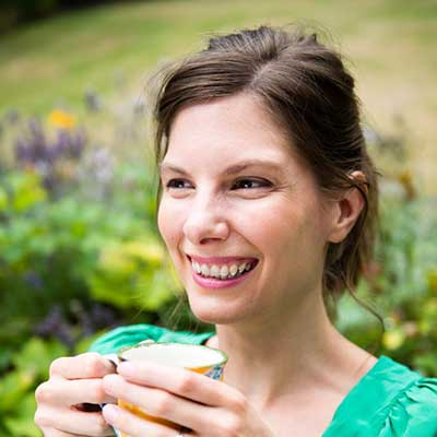 A person with brown hair holds a cup of coffee and smiles as they look into the distance