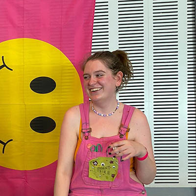 A smiling white woman with her hair pulled into a ponytail. She's wearing pink dungarees and standing in front of a pink flag with a yellow smiley face on it.