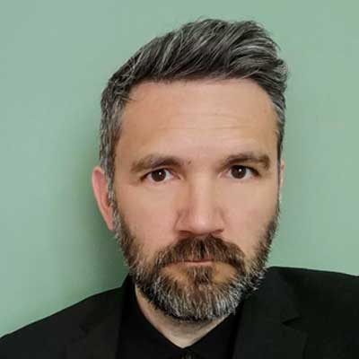A person with short dark grey hair and a beard looks at the camera