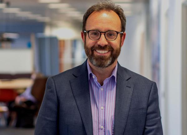 A man in a suit and glasses smiles warmly at the photographer, framed against a blurred office scene
