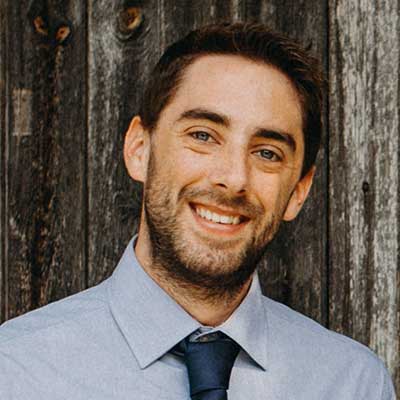 A person wearing a blue shirt and tie smiles at the camera