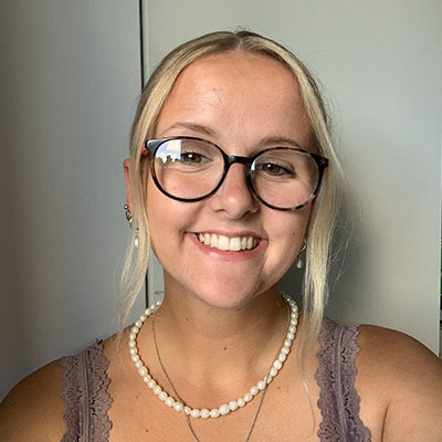 A student with blonde hair and glasses smiles at the camera