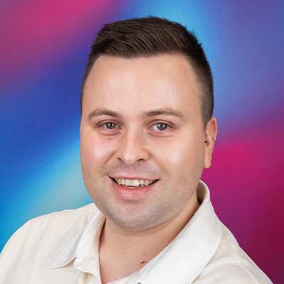 A person with short, dark hair smiles at the camera in front of a colourful background