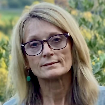 Image of person with glasses and long blonde hair smiling in front of foliage