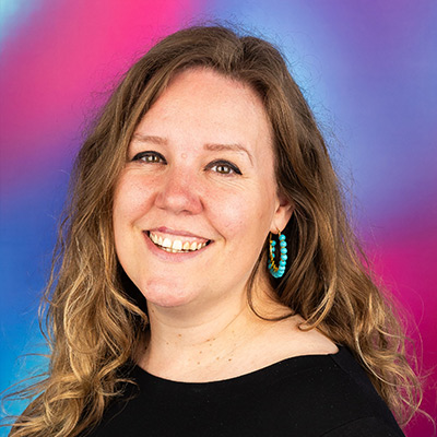 Headshot of person with long wavy light brown hair smiling in black top with turquoise ear-ring