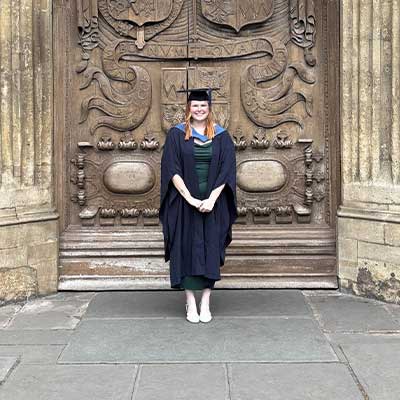 A person wearing navy graduation robes stands in front of a large ornate door outside Bath Abbey