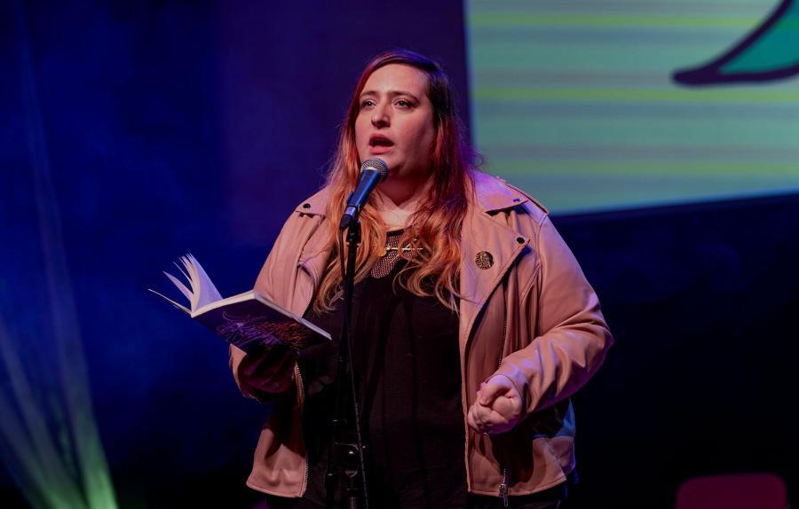 Woman on stage wearing a pink leather jacket and a confident expression holds an open book and speaks into a mic