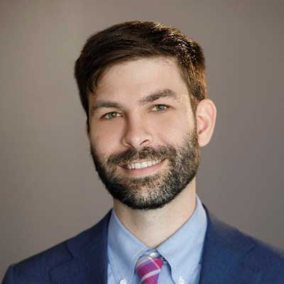 A person wearing a blue suit and tie smiles at the camera