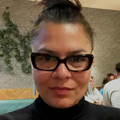 A woman with dark hair and glasses looks directly into the camera lens